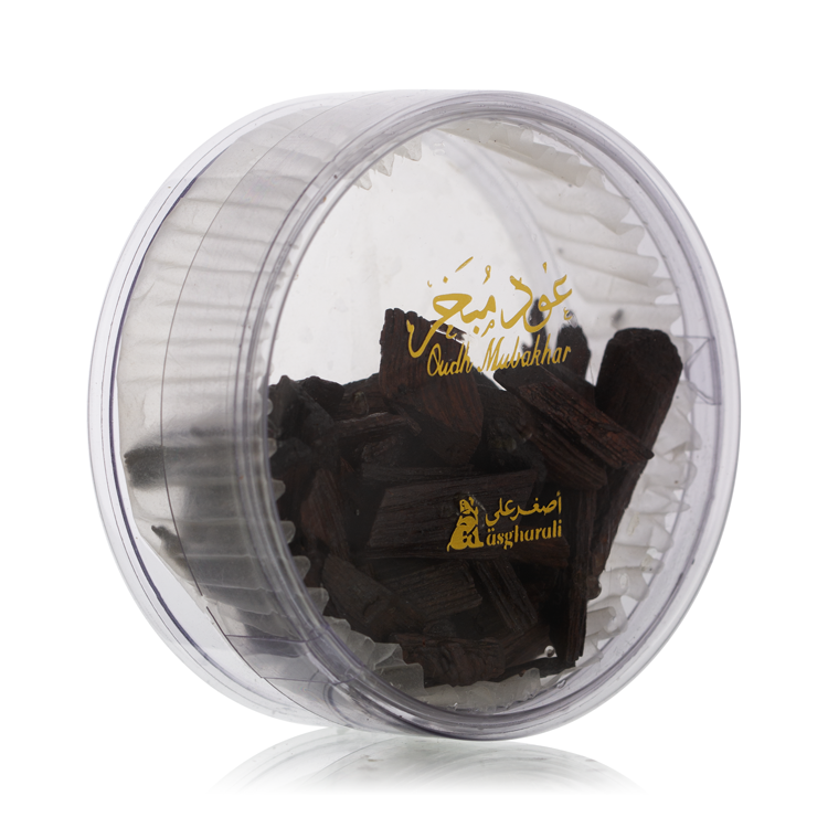 Close-up view of transparent round container filled with dark oud chips, labeled with golden Asgharali branding